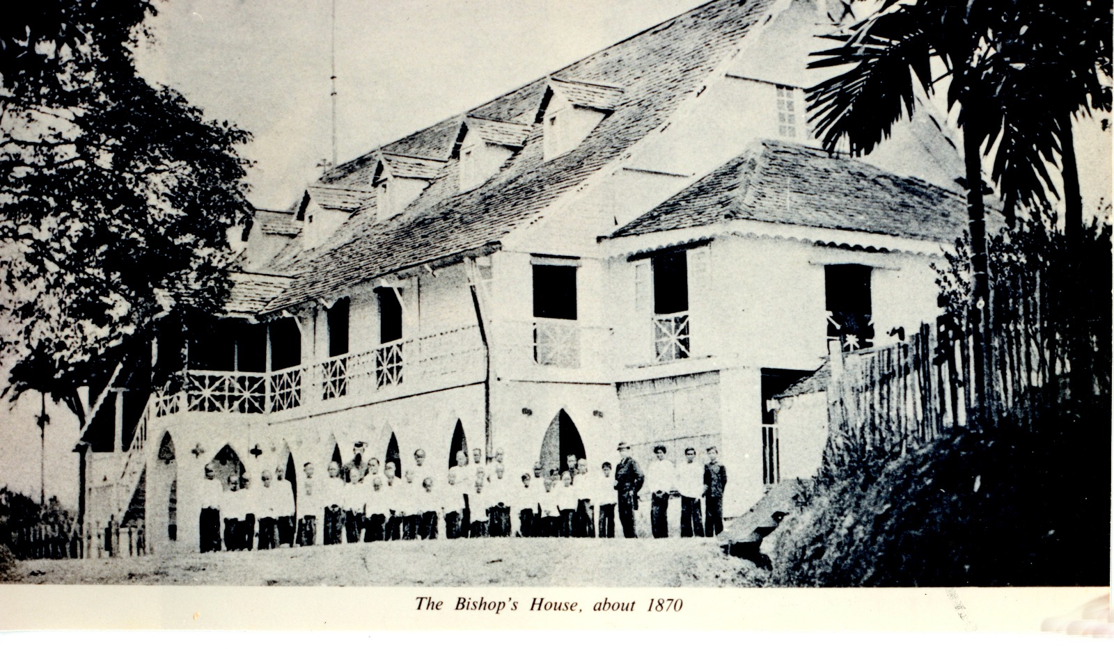Historical photograph from about 1870 of The Bishop's House in Kuching: a two-storey colonial building with Gothic arches and people gathered in front.